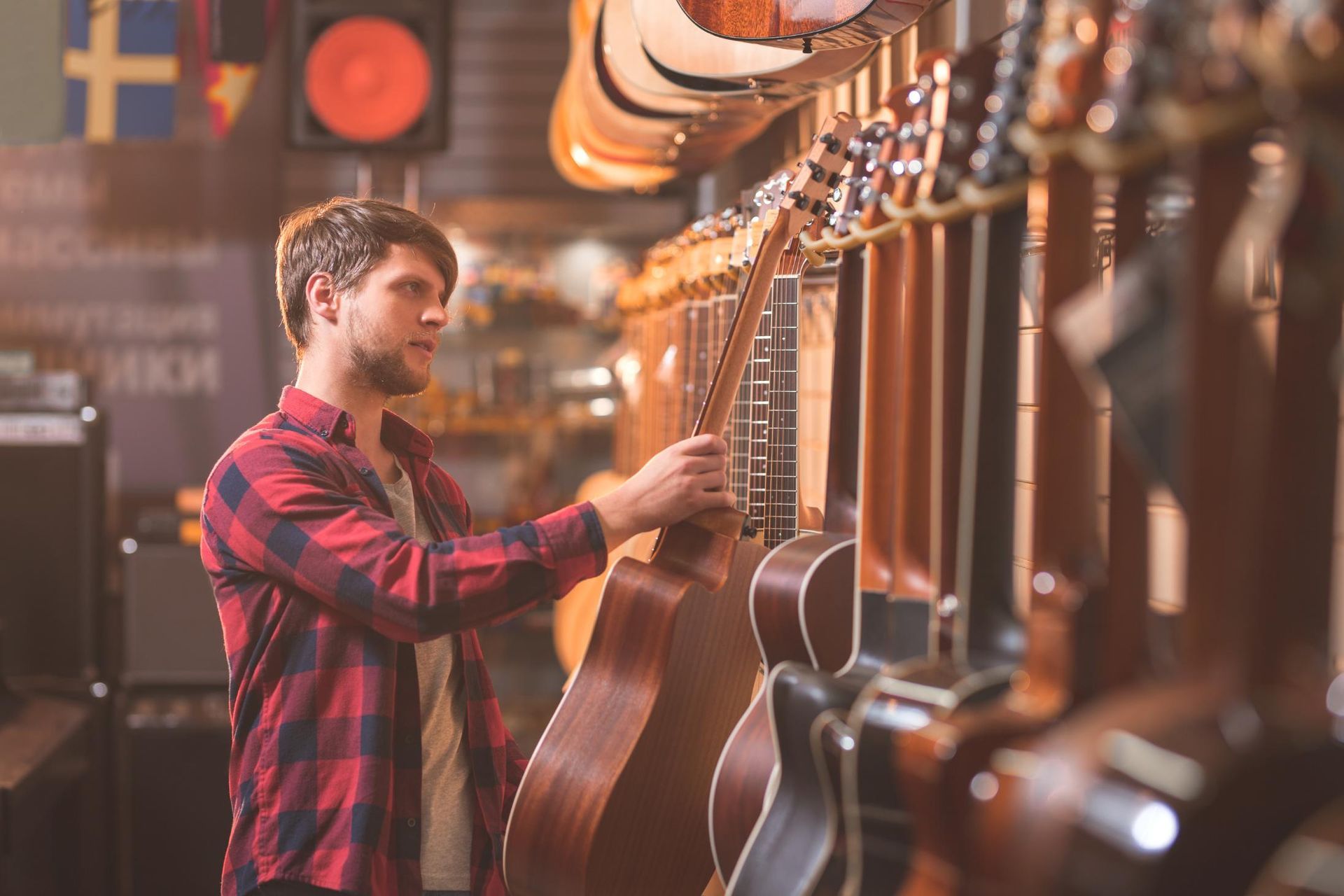 Man in a red plaid shirt examining acoustic guitars in a warmly lit music shop
