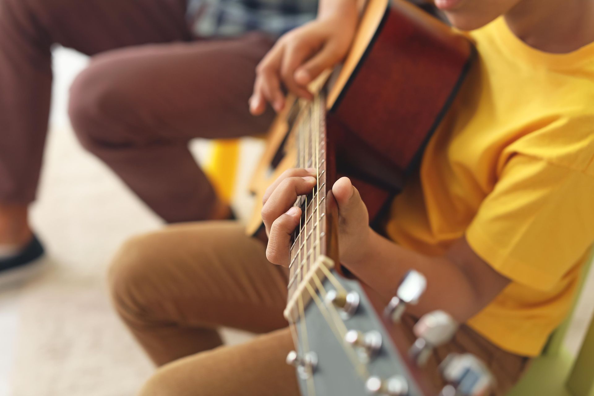 Close-up of hands playing an acoustic guitar, with a person in a yellow shirt seated nearby