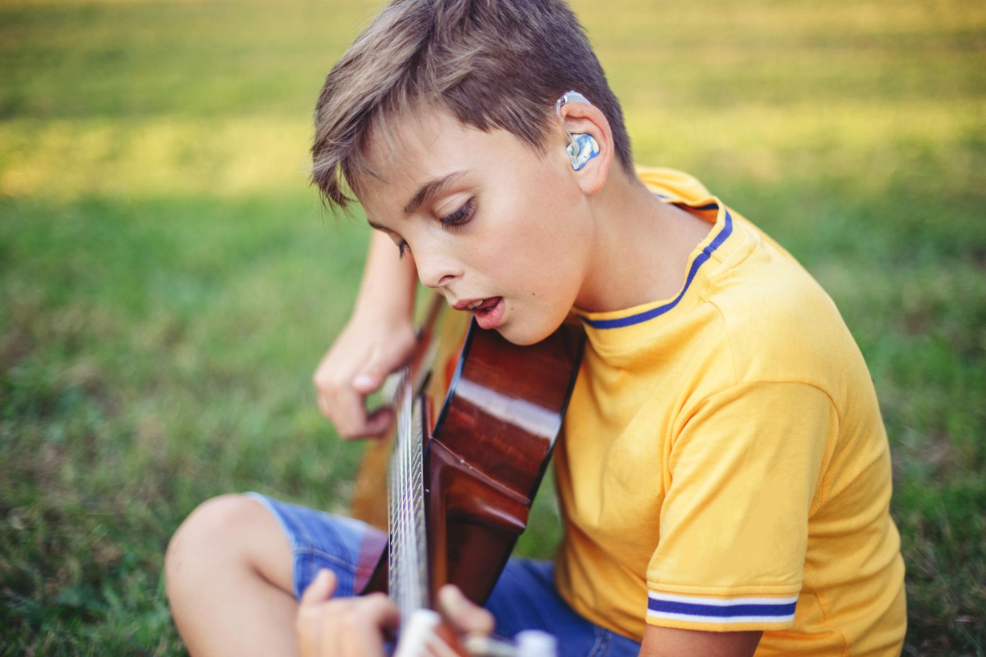Child in a yellow shirt playing an acoustic guitar outdoors on grass