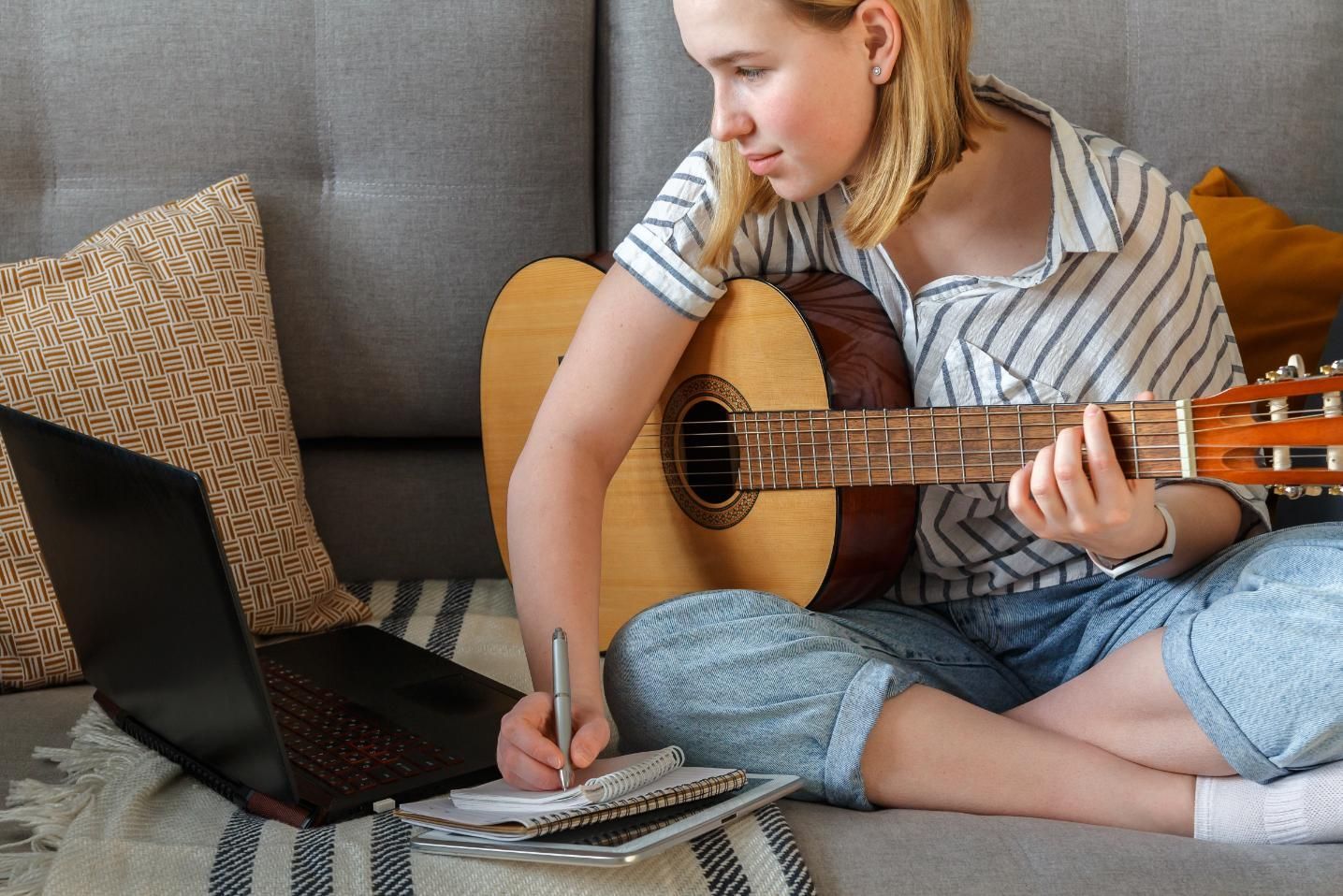 Person sitting on a sofa, writing in a notebook with a laptop and guitar beside them