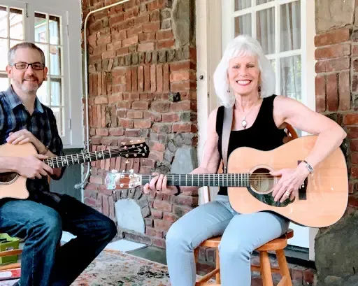 Two people seated on a porch playing acoustic guitars beside a brick wall and window