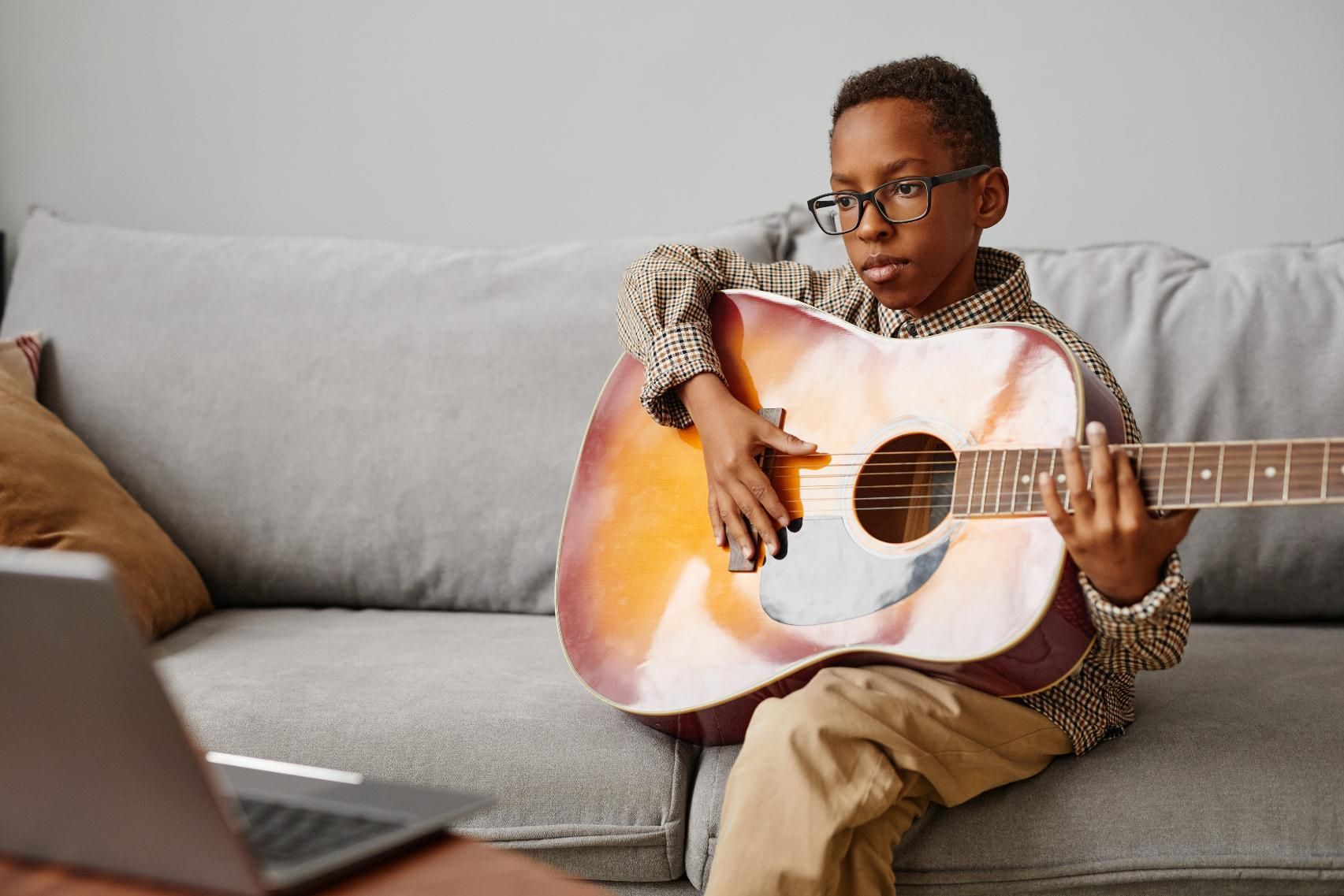 Person playing acoustic guitar on a gray couch beside a laptop in a living room