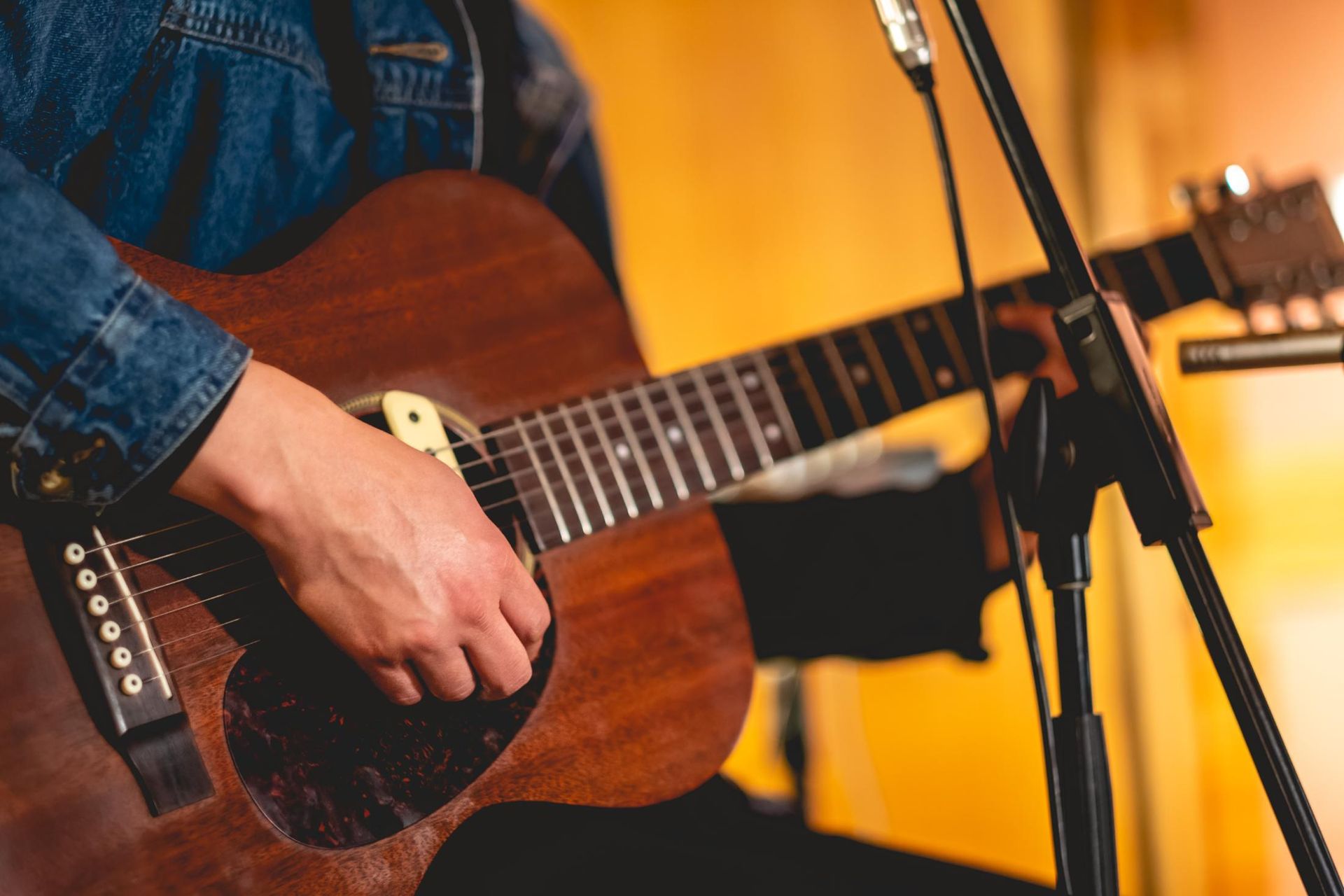 Hand playing acoustic guitar on stage with warm orange lighting and a microphone stand nearby