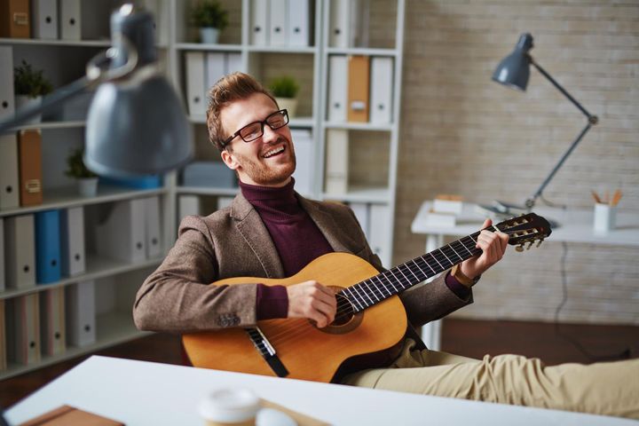 Smiling man playing acoustic guitar in a cozy office with shelves and desk lamps