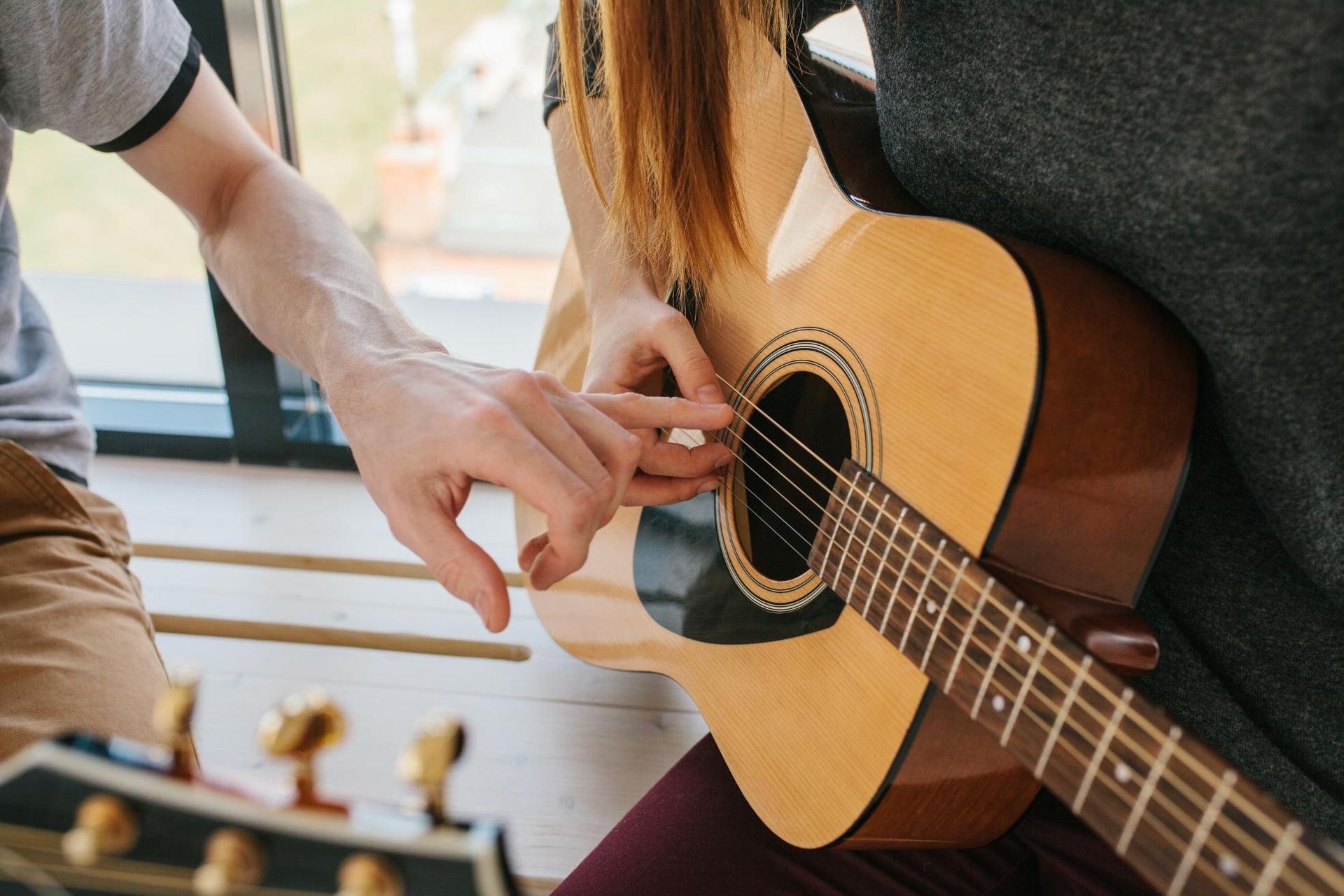 Hands adjusting a guitar string on an acoustic guitar beside a seated player outdoors