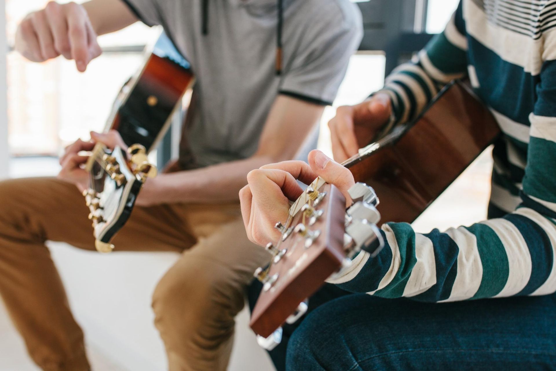 Two people strumming acoustic guitars indoors, one in a striped sweater.