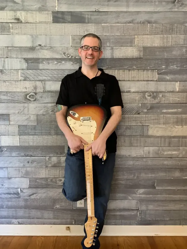 Smiling person in a black shirt and jeans leaning on a guitar against a gray wood wall