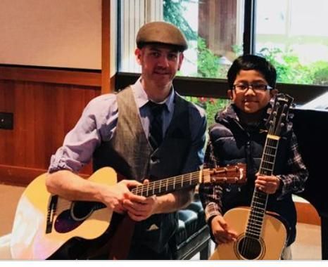 Two people smiling and holding acoustic guitars in a wood-paneled room near a window