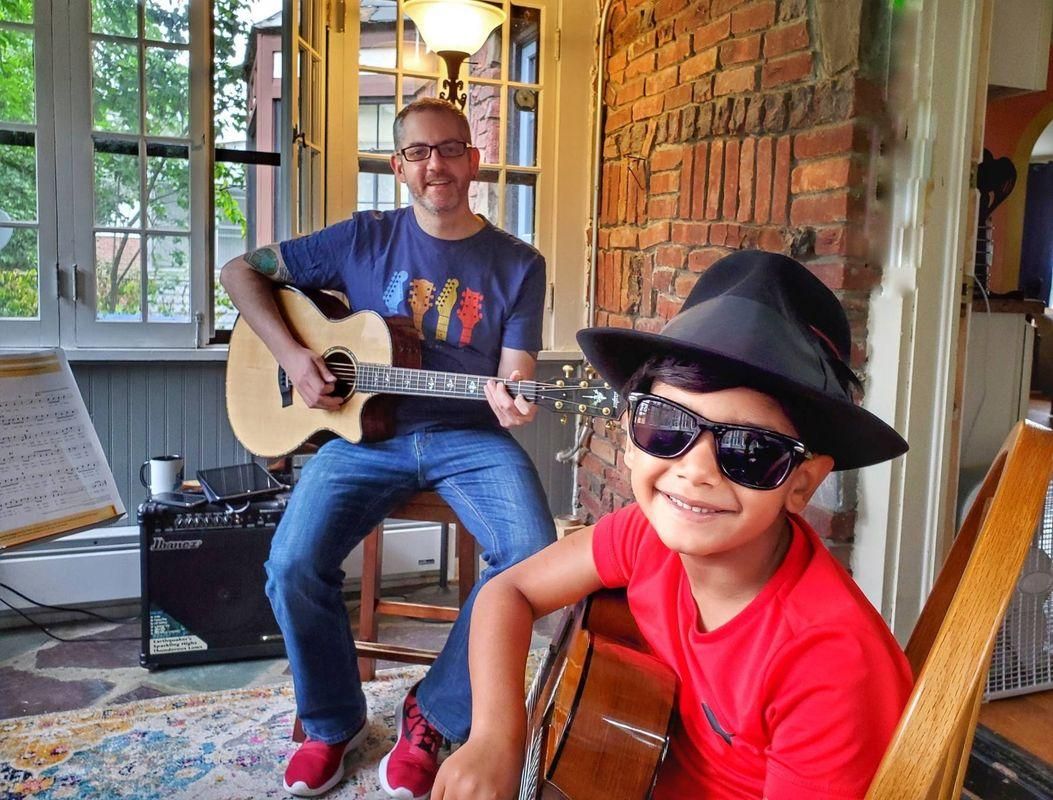 Two people playing acoustic guitars on a porch, one smiling in sunglasses and a hat.
