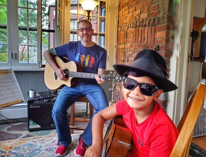 Two people playing acoustic guitars on a porch, one wearing a hat and sunglasses, smiling.