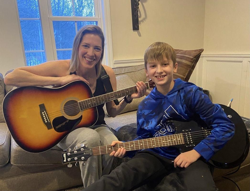 Two people smiling with acoustic guitars on a couch in a living room.