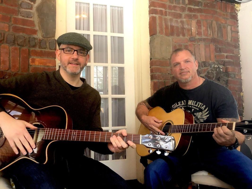 Two men seated with guitars in front of a brick wall and window, smiling in a casual indoor setting