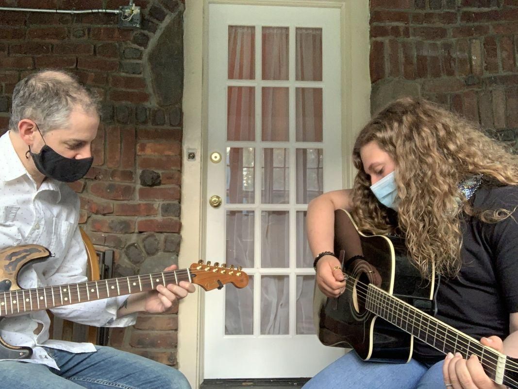 Two masked musicians seated outdoors, playing guitars in front of a brick wall and white door.