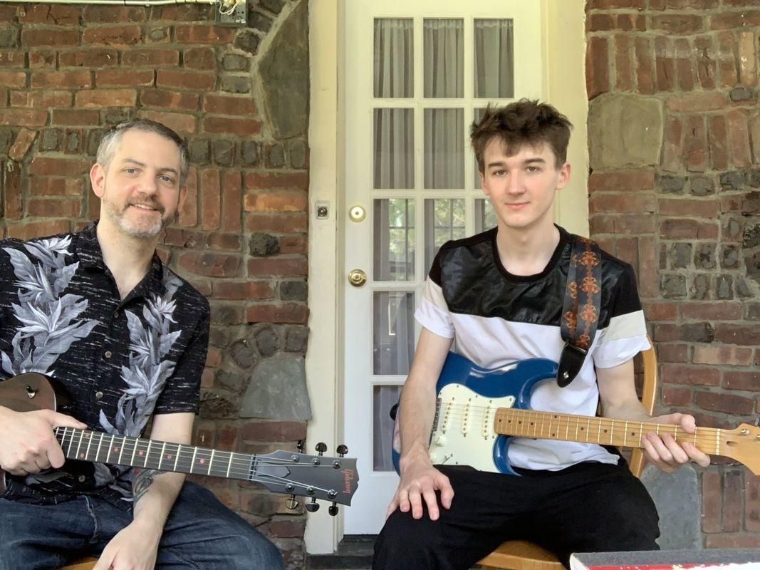 Two people seated with electric guitars on a brick porch in front of a white door