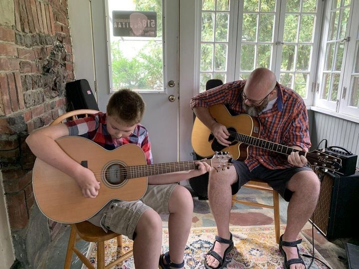 Two people playing acoustic guitars on a porch, seated near glass doors and windows.