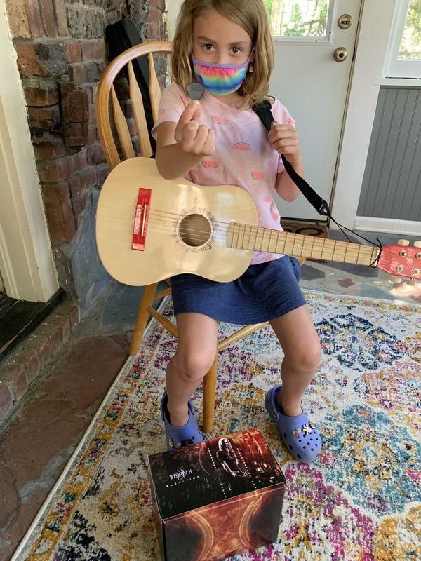 Child sitting indoors with a small guitar on a decorated chair, wearing a rainbow face mask and blue clogs.
