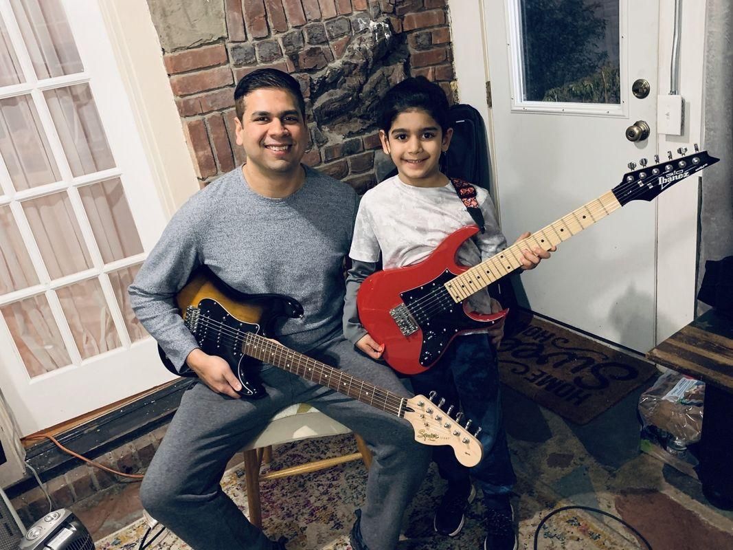 Two people holding electric guitars, smiling indoors near a brick wall and front door.