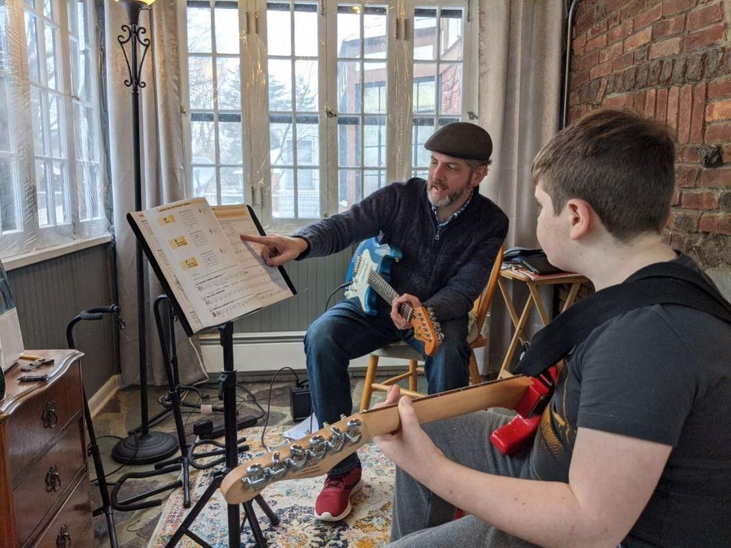 Two musicians rehearsing in a bright room, one reading sheet music while holding guitars.
