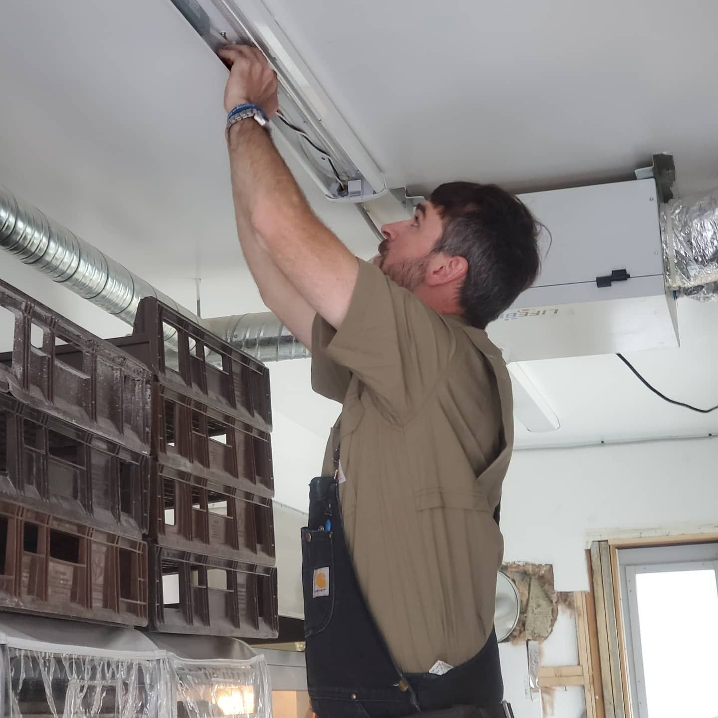 Technician adjusting a ceiling-mounted unit in a room with stacked storage crates and exposed wiring