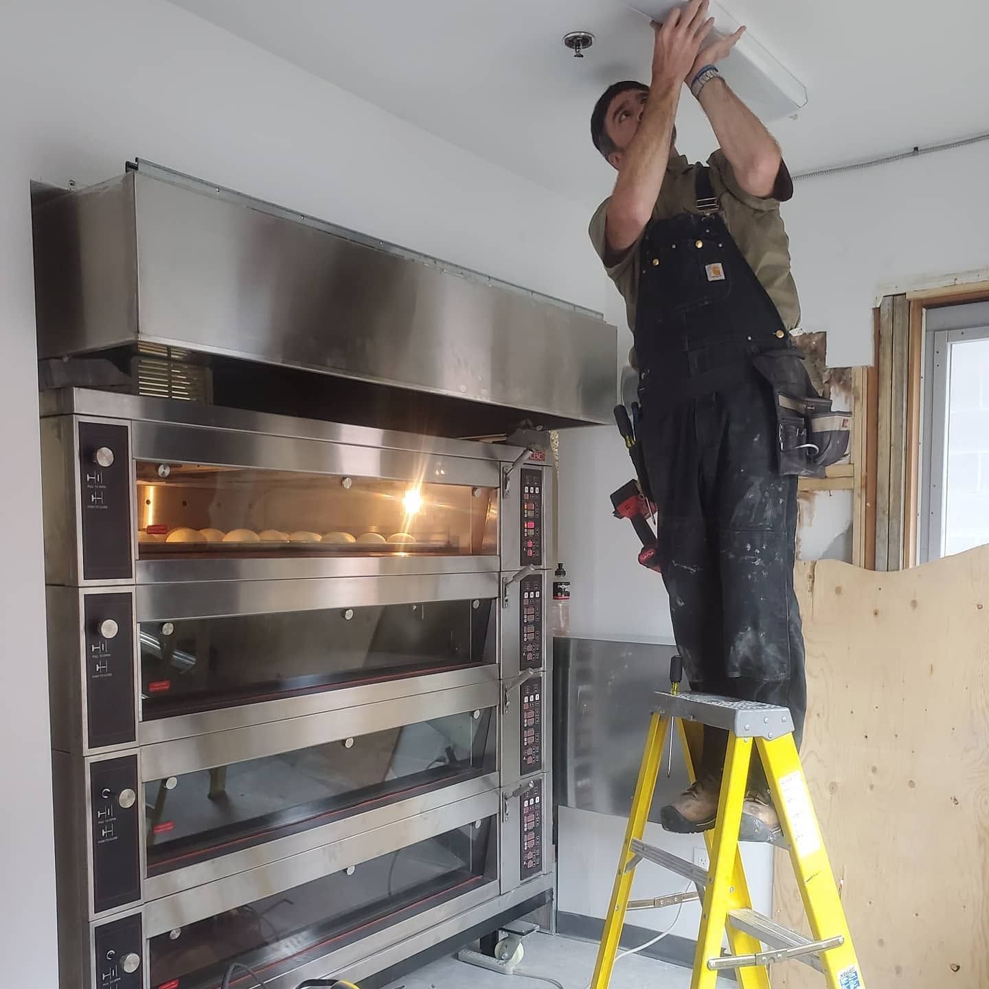 Person on a yellow ladder installing a ceiling light in a kitchen with a large stainless-steel oven