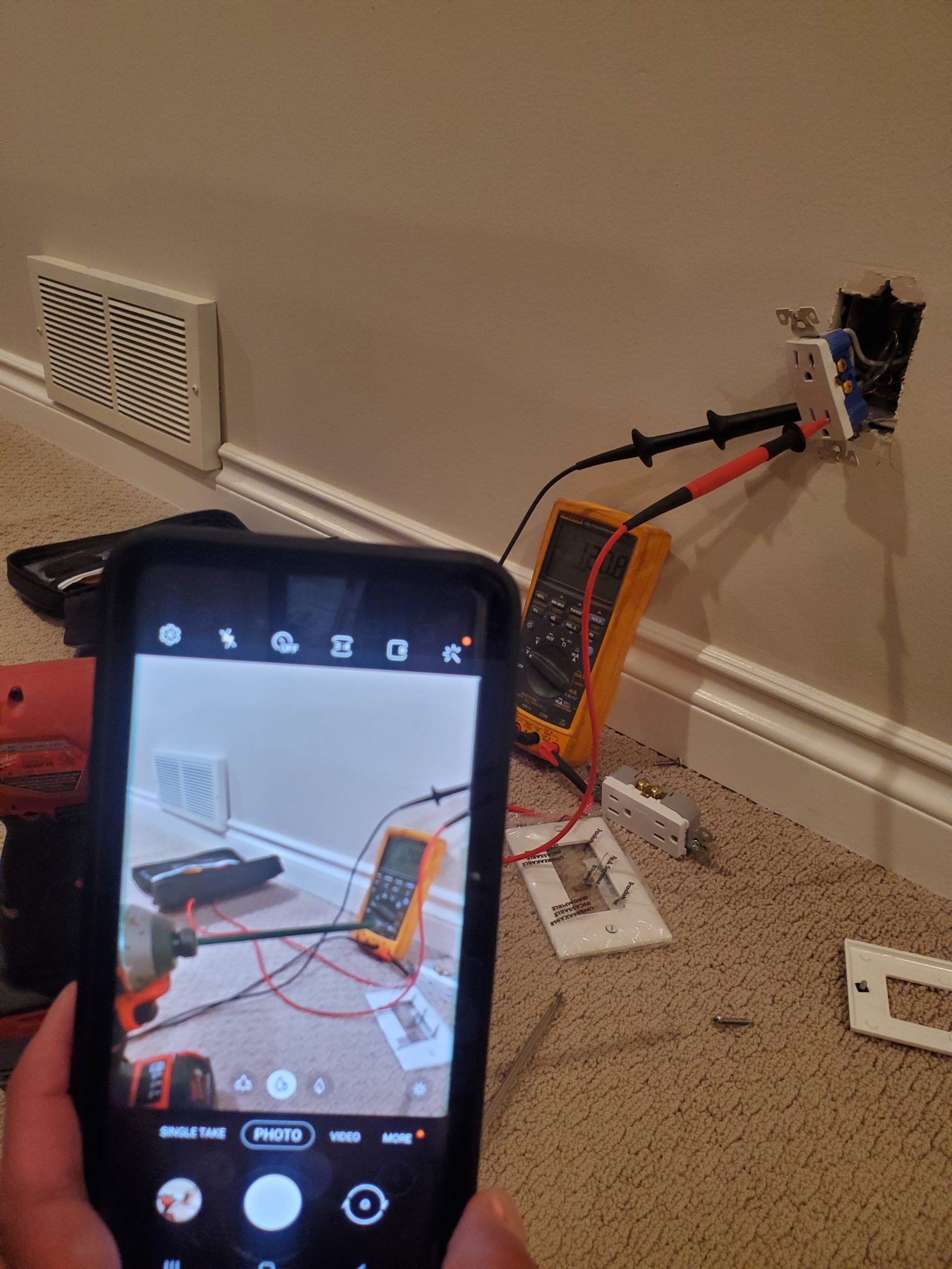 Hand holding a phone photographing tangled wires and a tilted orange device against a wall by a vent