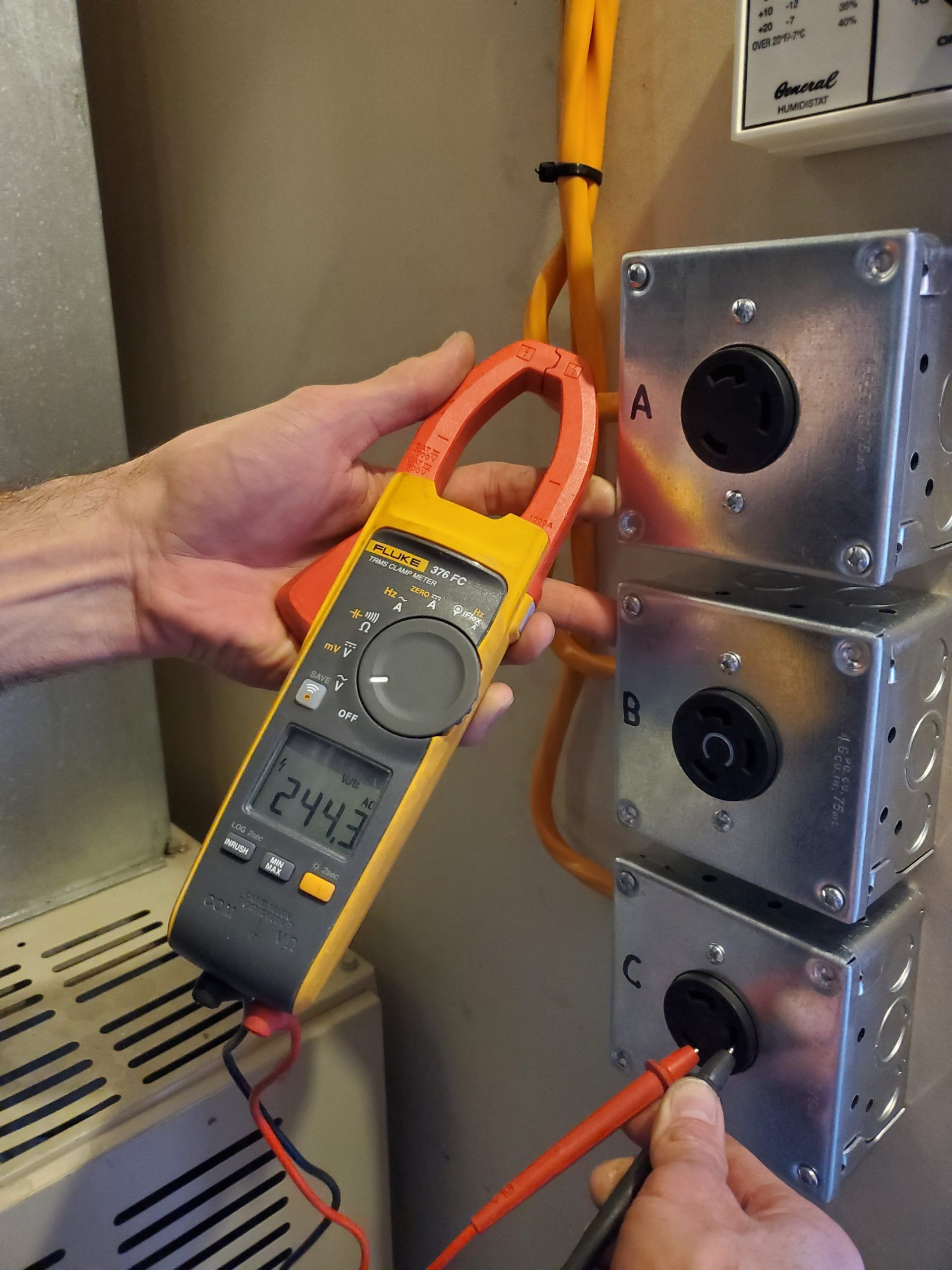 Handheld digital multimeter testing three stacked electrical boxes with orange and red leads
