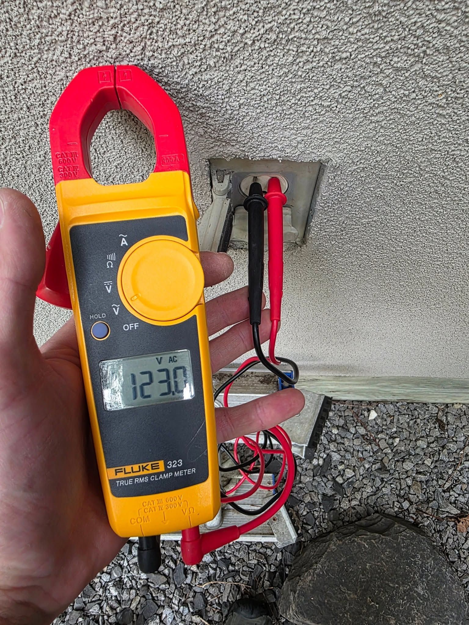 Handheld clamp meter showing 123.6, held near exposed electrical wires against a wall.