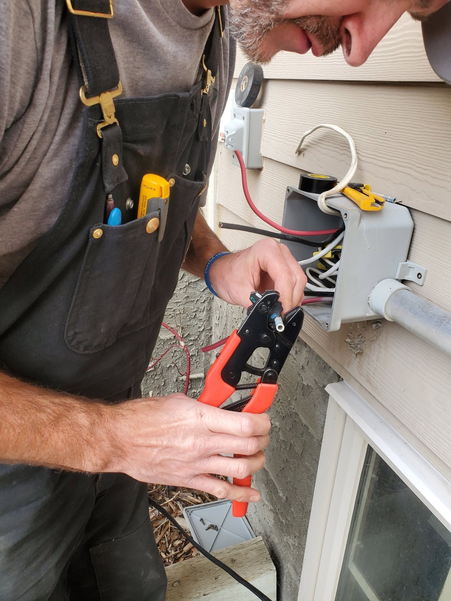 Electrician using a red wire cutter to work on outdoor house wiring beside a window