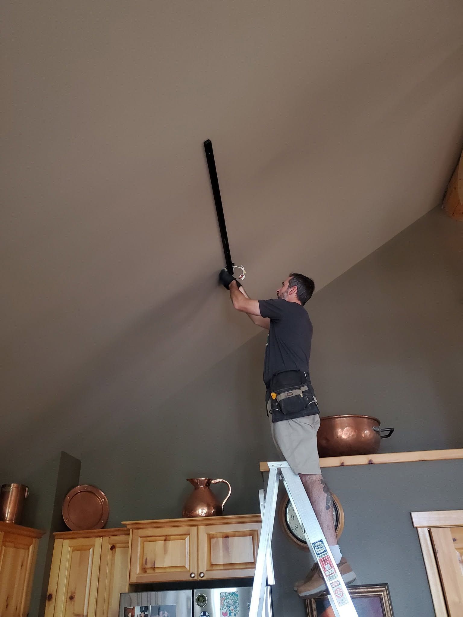 Person on a ladder reaching a long pole toward a high ceiling in a kitchen-like room