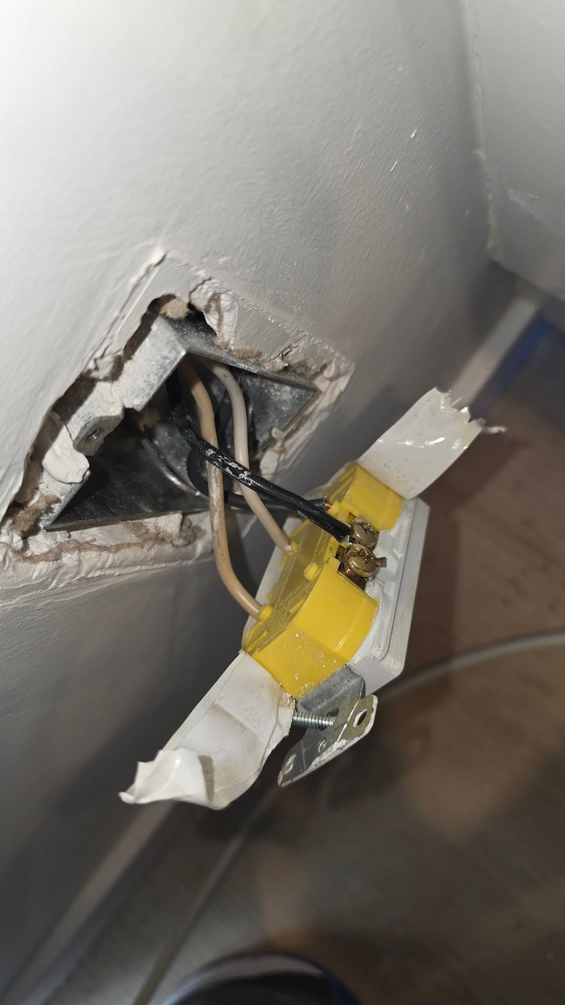 Damaged wall outlet with exposed black wires and yellow tape hanging out of a hole in a white wall