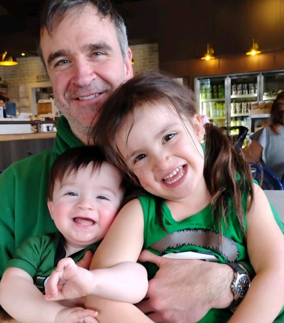 Smiling man with two children in green shirts, hugging closely indoors at a restaurant or café