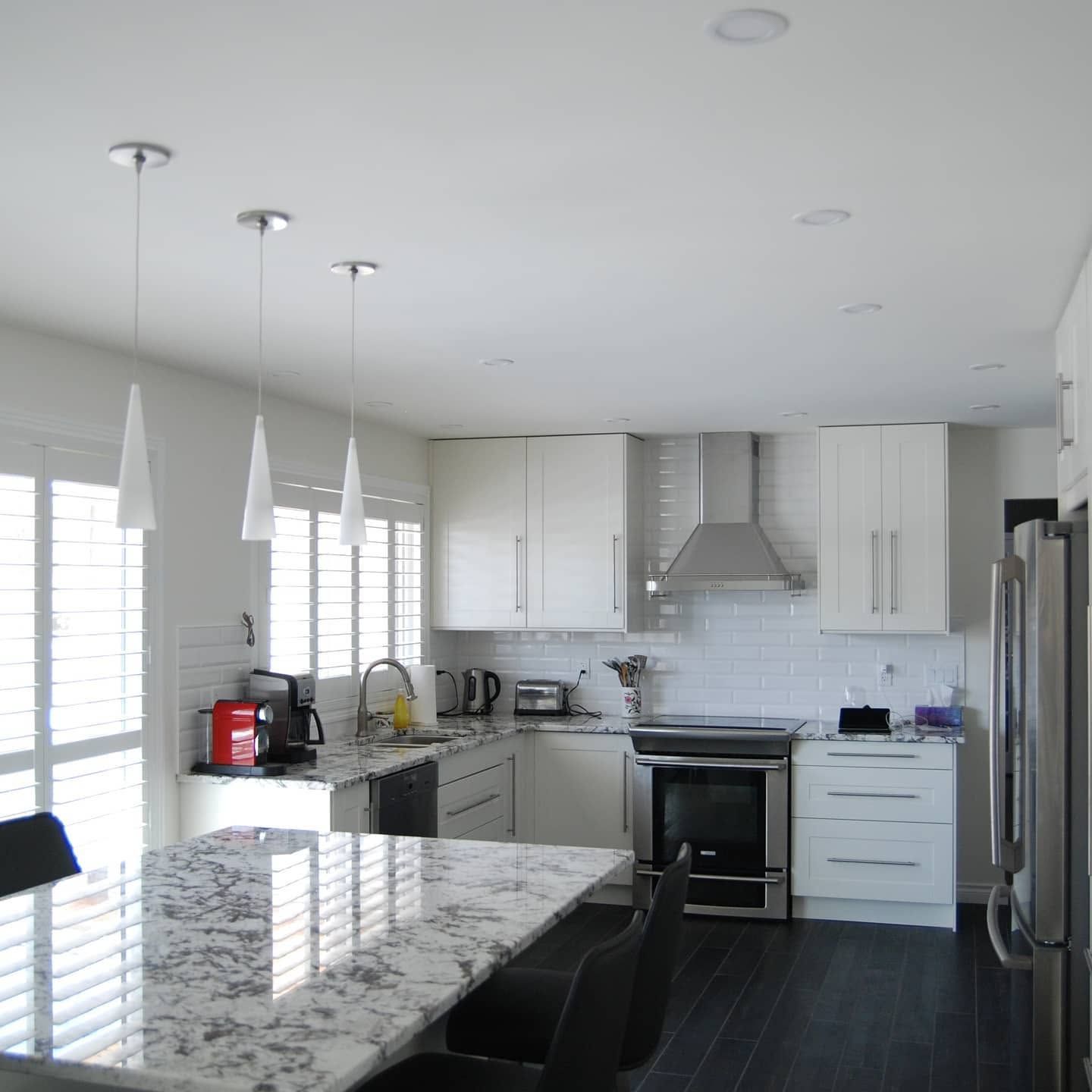 Modern white kitchen with granite island, pendant lights, stainless appliances, and dark wood floor.