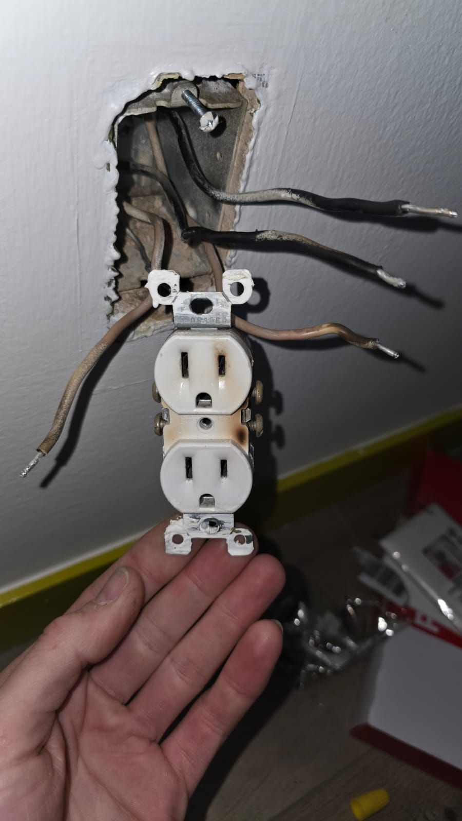 Open wall outlet being rewired with exposed black and brown wires and a hand holding the plate