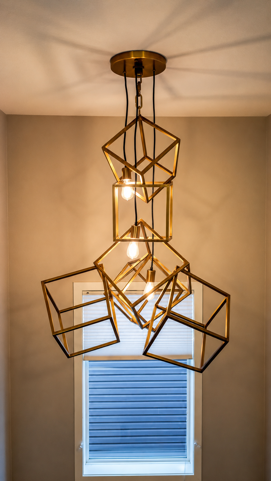 Geometric metal pendant chandelier with three lit bulbs hanging above a window in a beige room