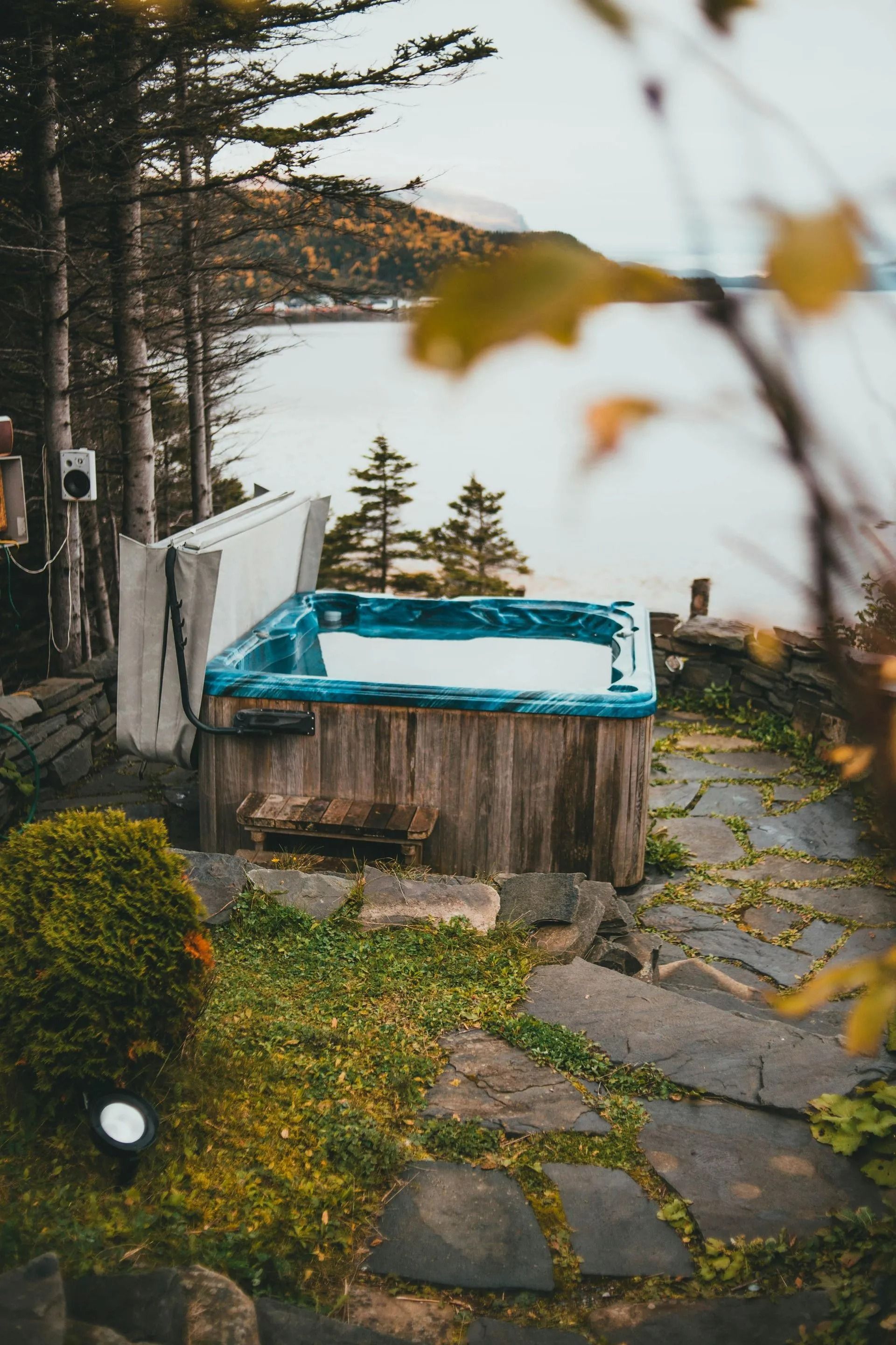 Outdoor hot tub beside a lake, framed by trees and stone steps, with a forested shoreline beyond.