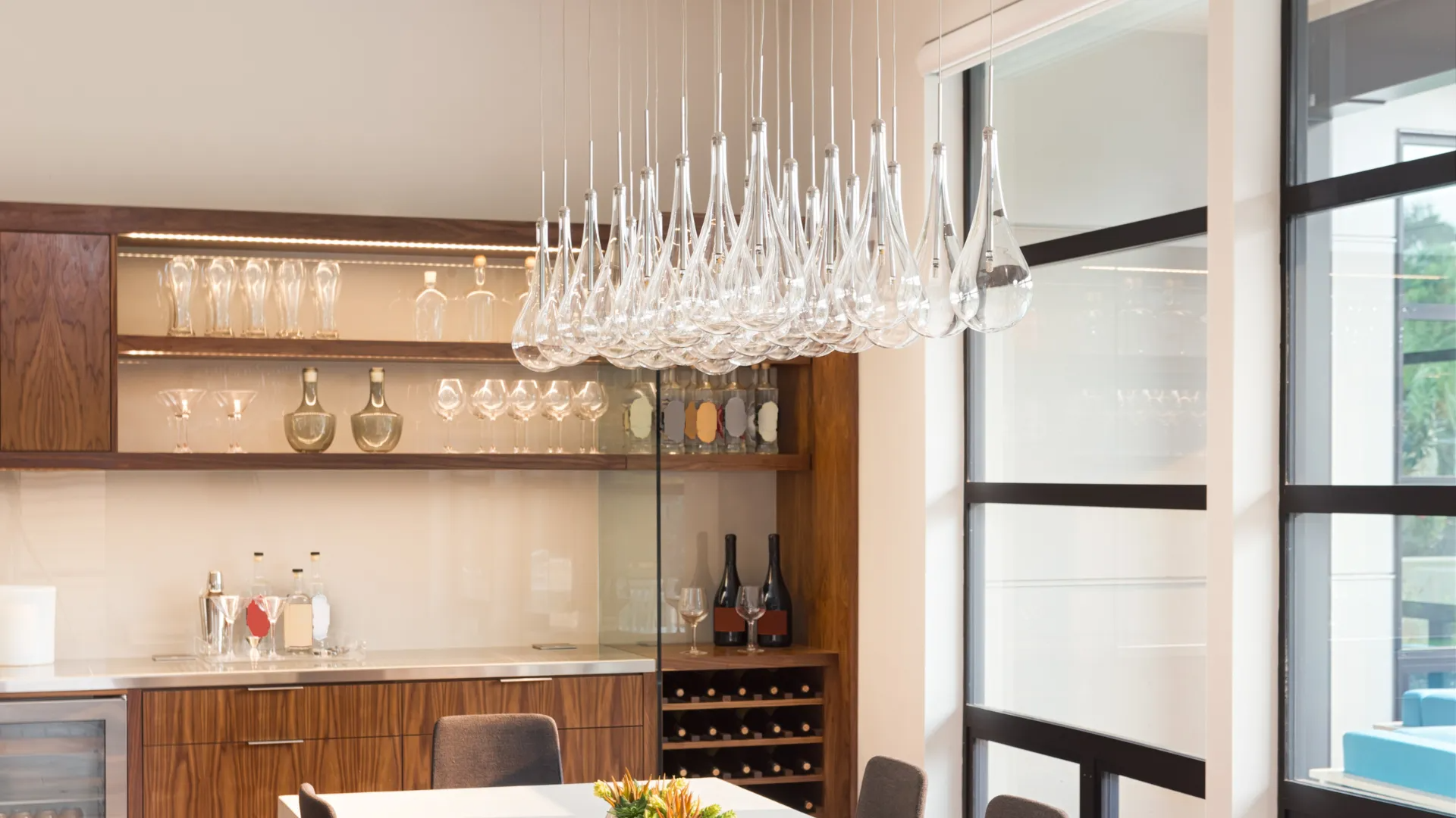 Modern kitchen with wood cabinets, glassware, and a row of hanging pendant lights by a window.