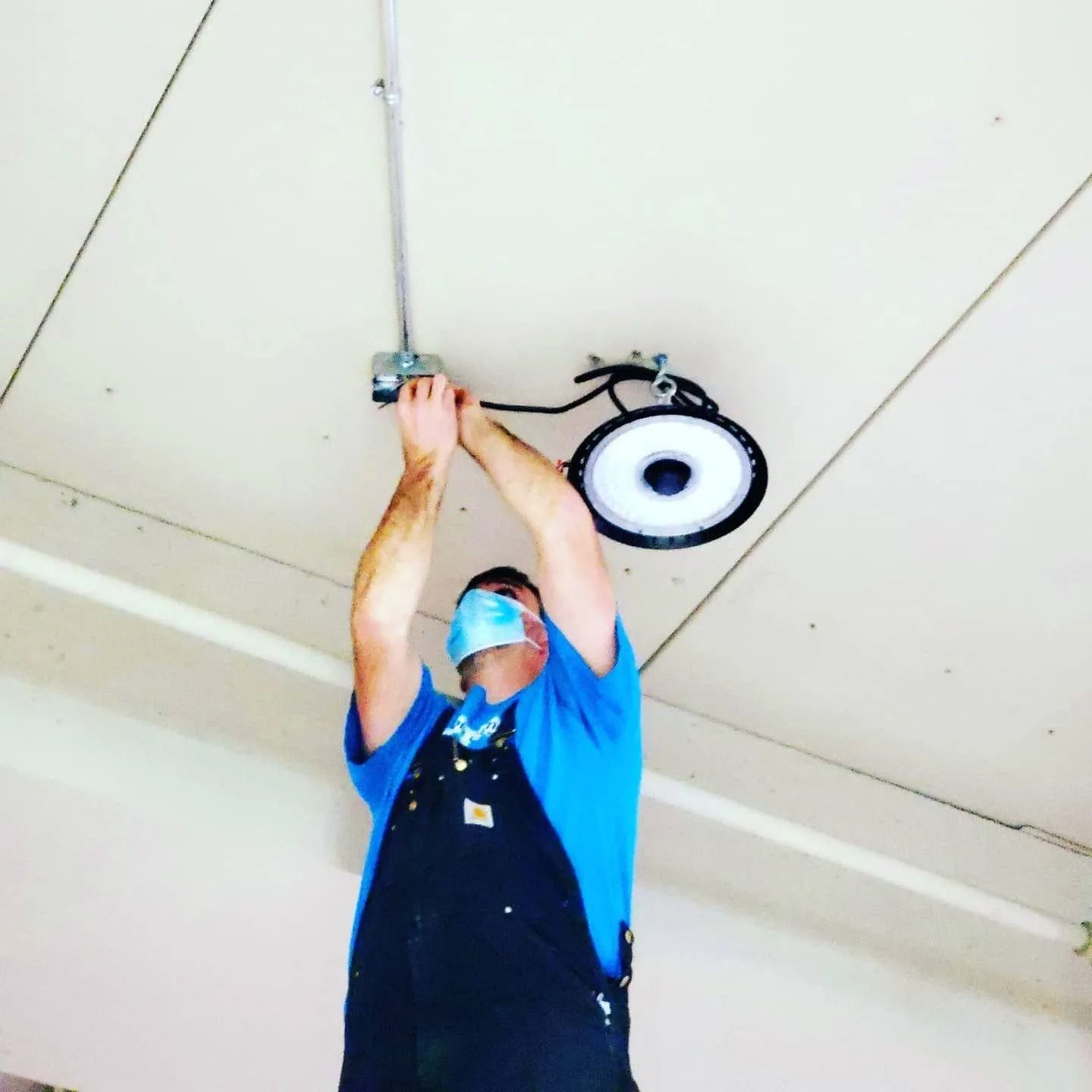 Worker in blue shirt adjusting a ceiling light fixture while standing on a ladder, wearing a face mask