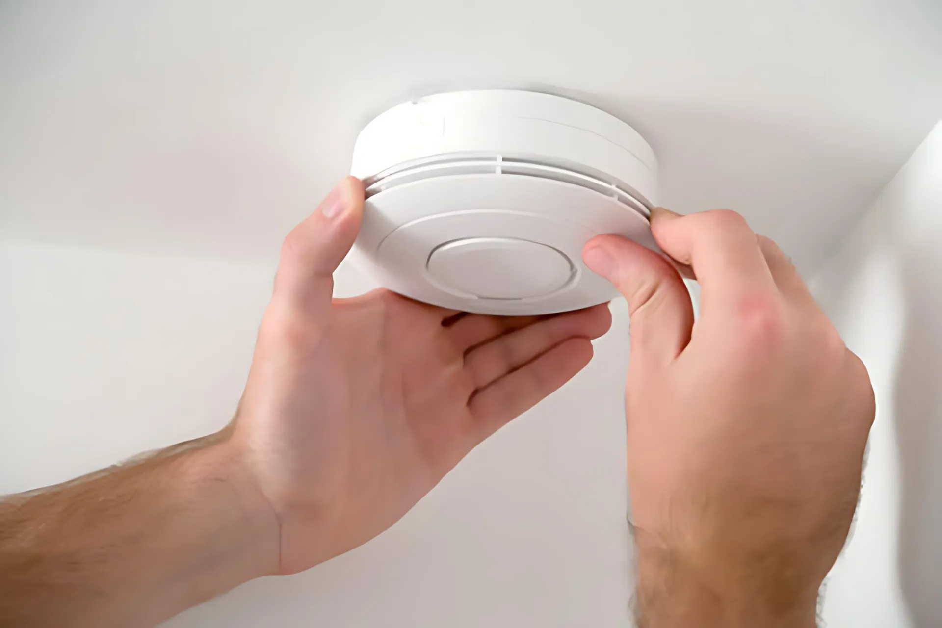 Hands installing a white smoke detector on a ceiling