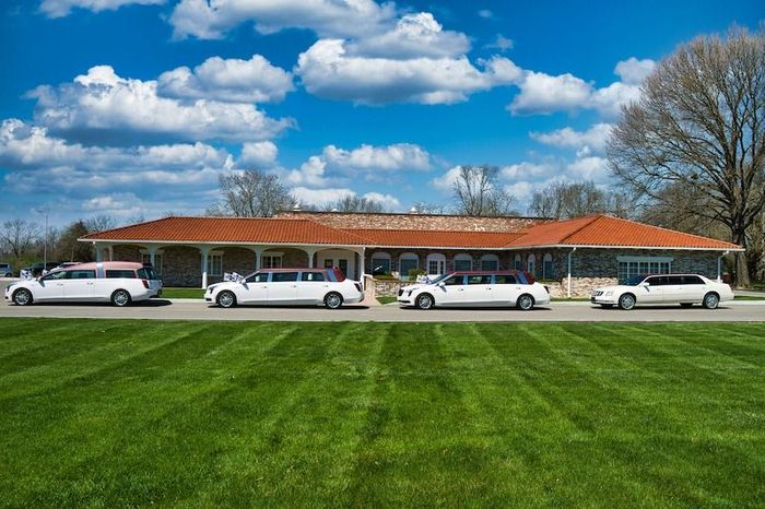 Four white limousines parked in front of a building with an orange roof under a bright blue sky with clouds.