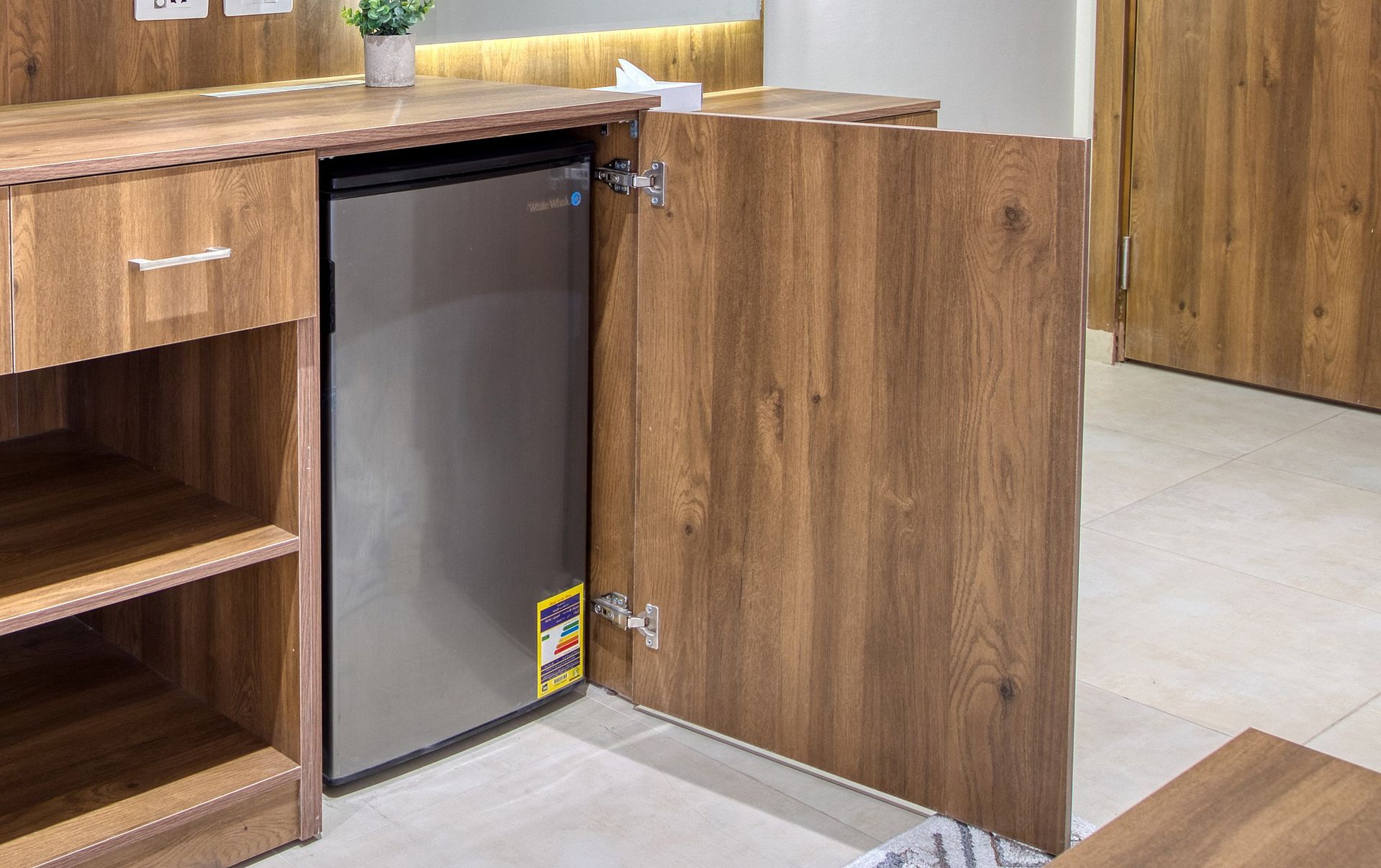 A small refrigerator is sitting under a wooden cabinet in a room.