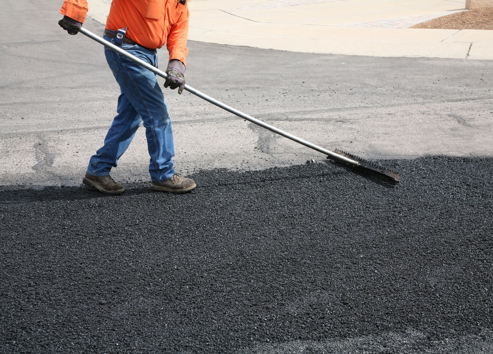 Person in work attire using a long-handled tool to smooth fresh asphalt on a roadway.