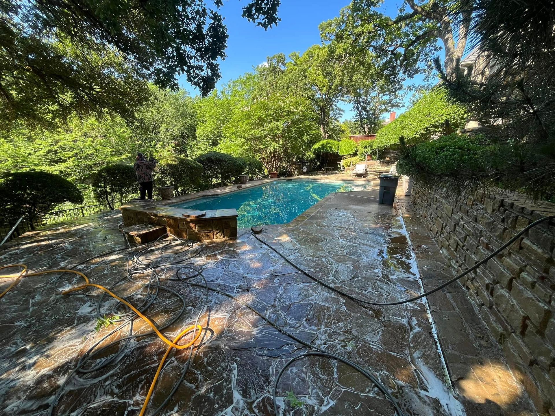 Person pressure washing a stone patio near a blue pool surrounded by trees.