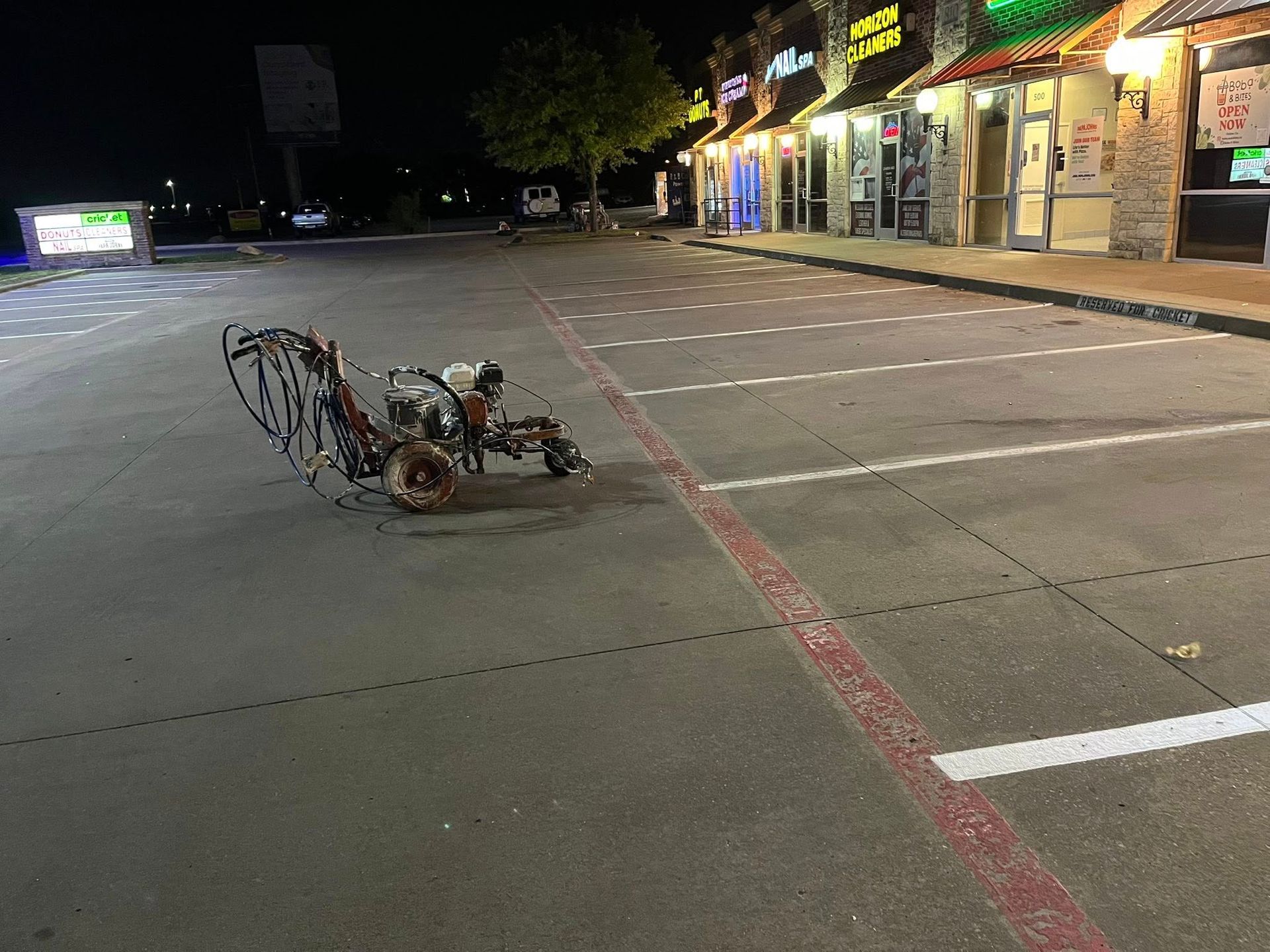 Rusty, metal sculpture in a nearly empty parking lot at night, in front of a row of illuminated storefronts.