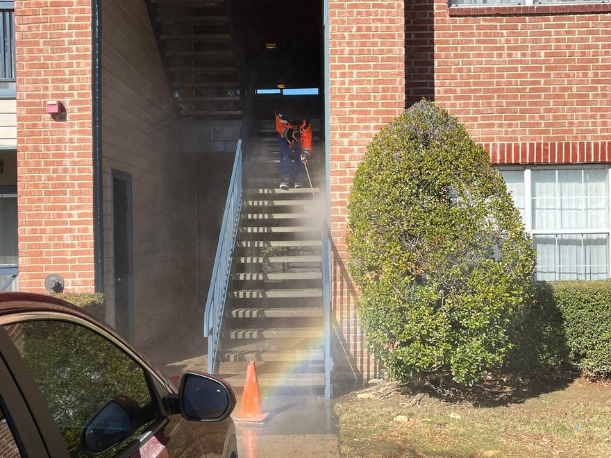 People power washing outdoor stairs, creating a rainbow effect, near a brick building and car.