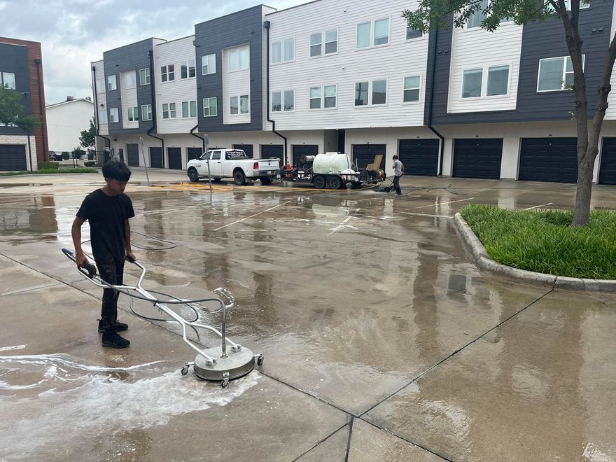 Person power washing a concrete surface in front of apartment buildings; wet concrete, cloudy sky.