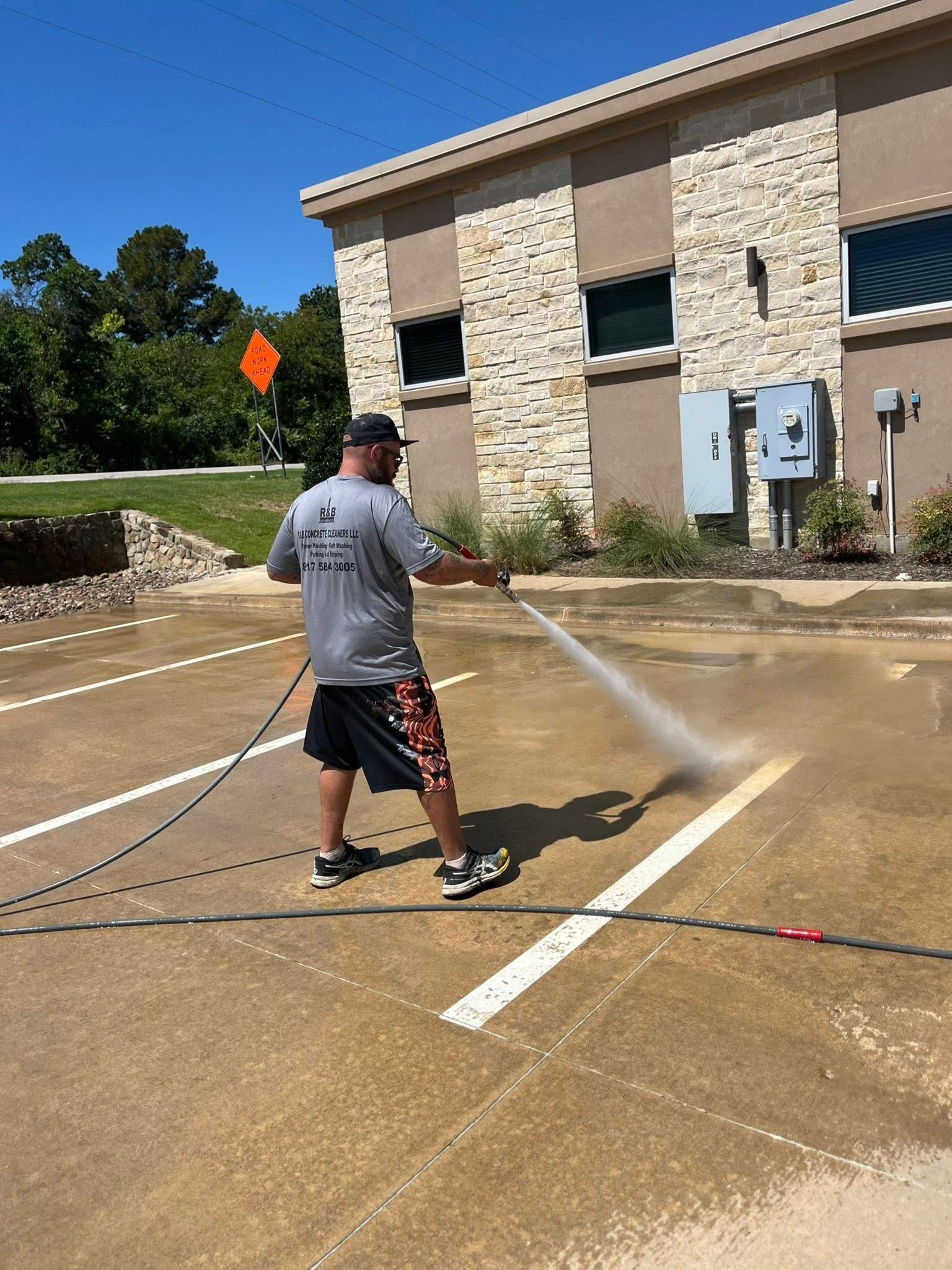 Man power washing a parking space line in front of a beige building on a sunny day.