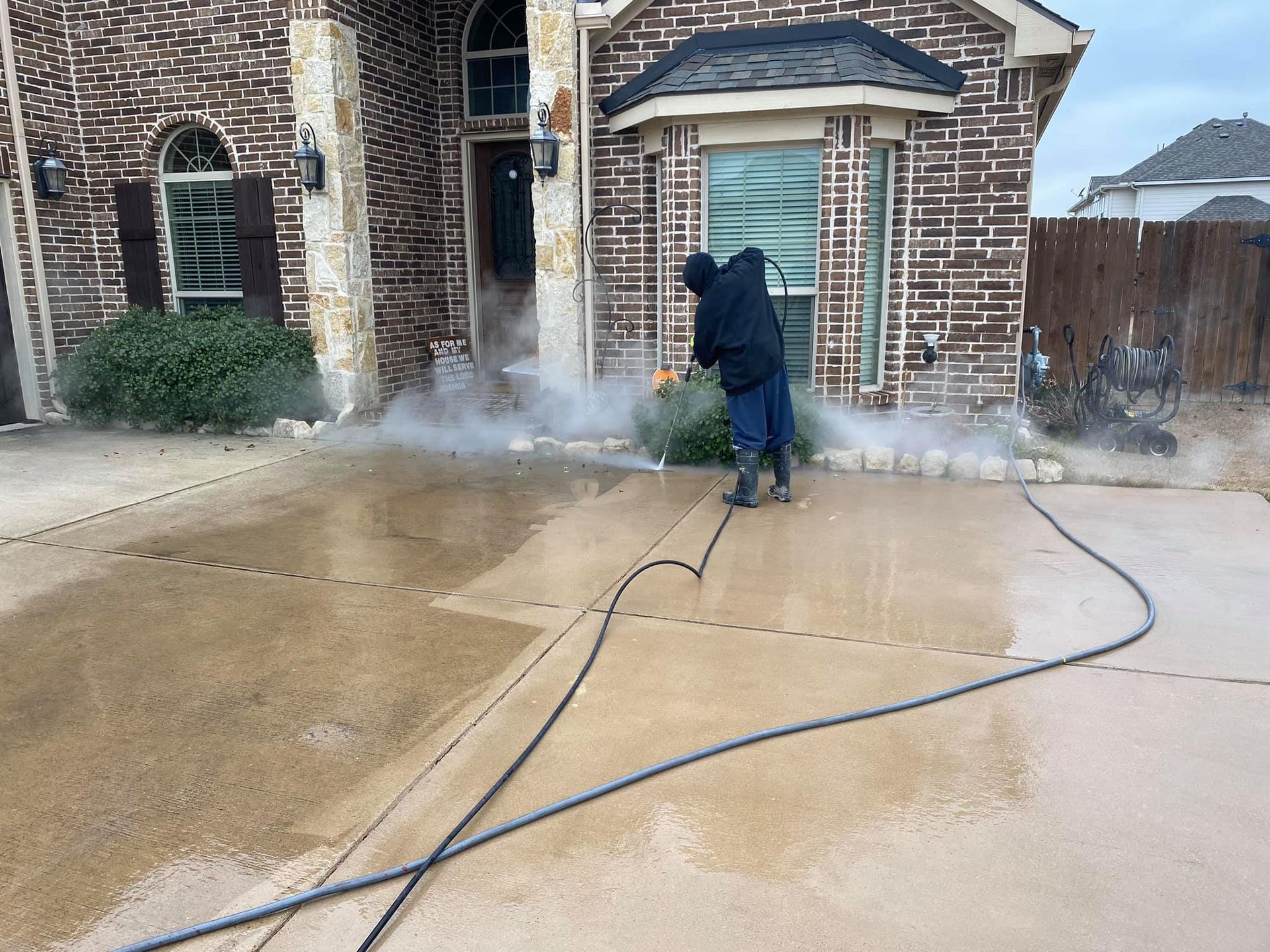 Person pressure washing a concrete driveway in front of a brick house.