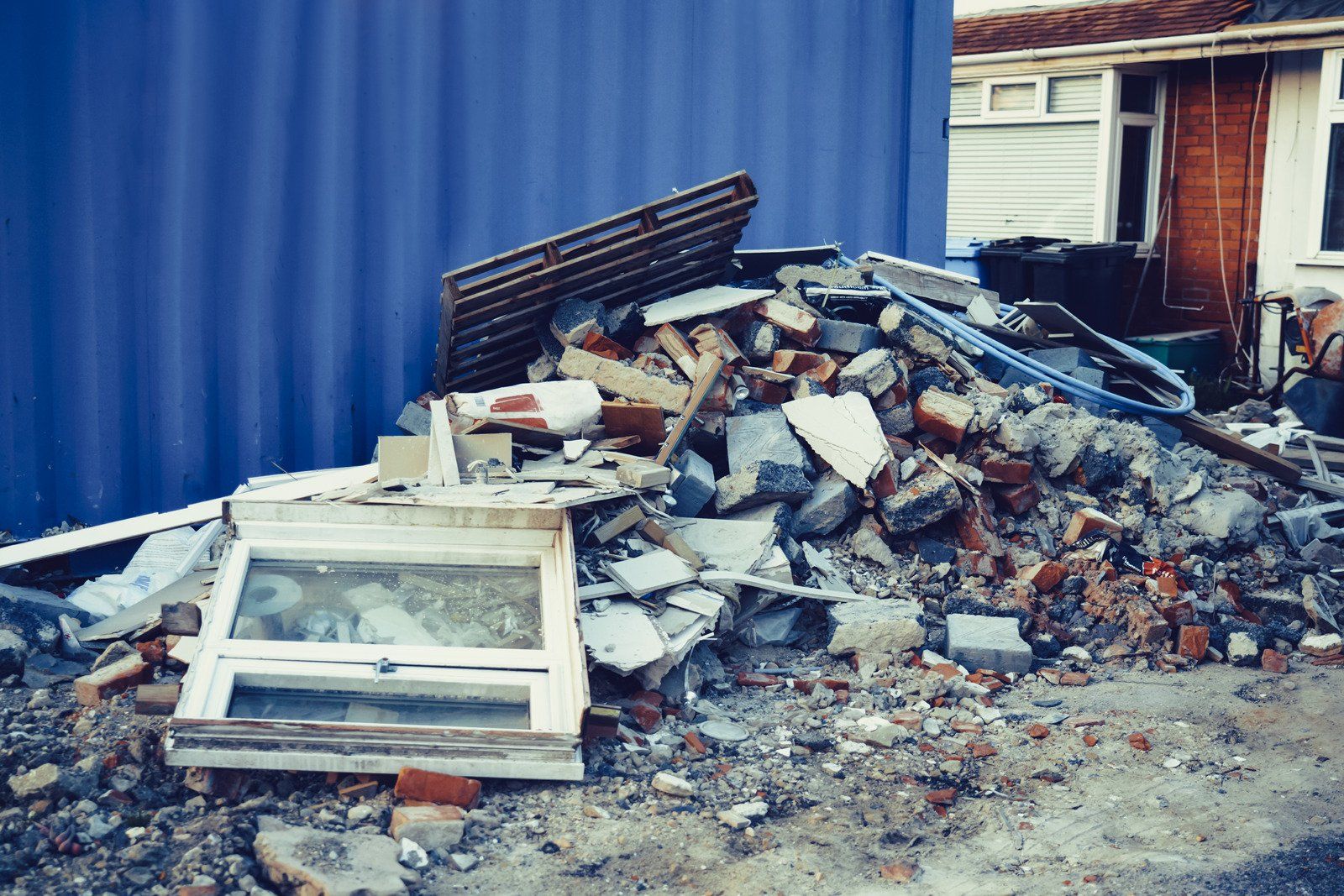 Pile of demolition debris, including a window, next to a blue wall and a building with a brick facade.