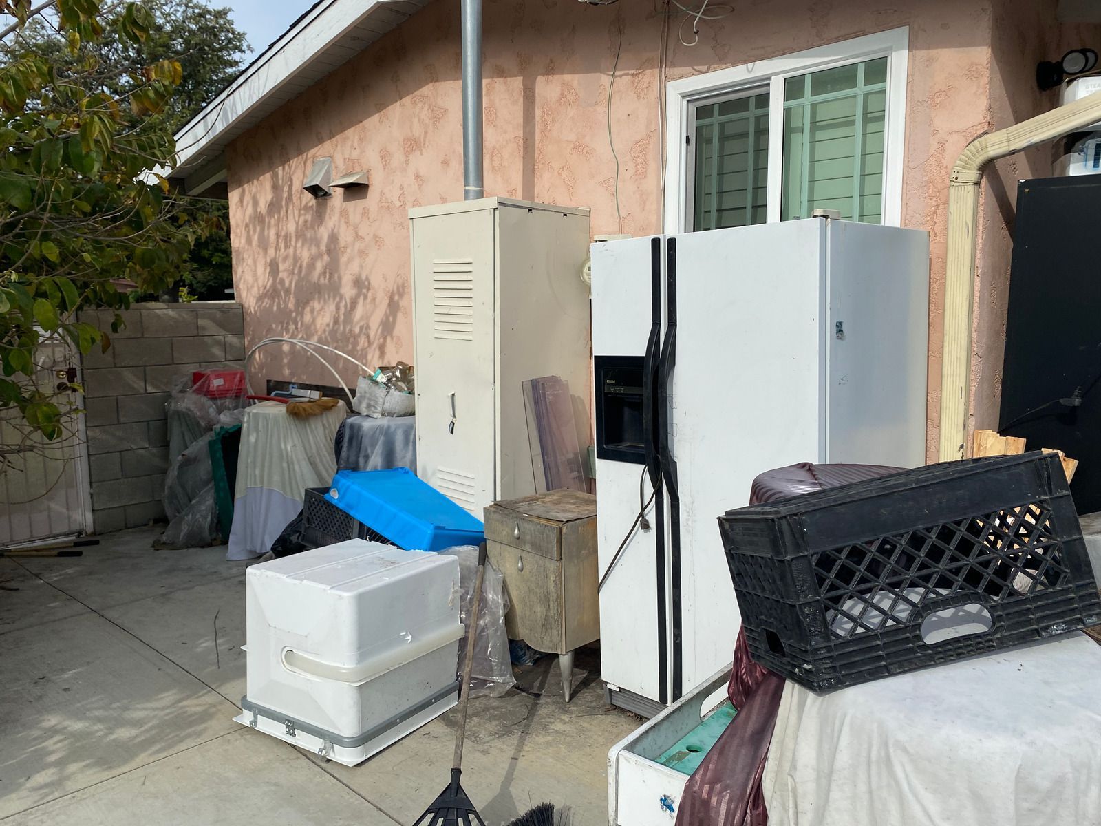 Backyard cluttered with appliances, boxes, and debris; pink stucco wall, window, and gray concrete.