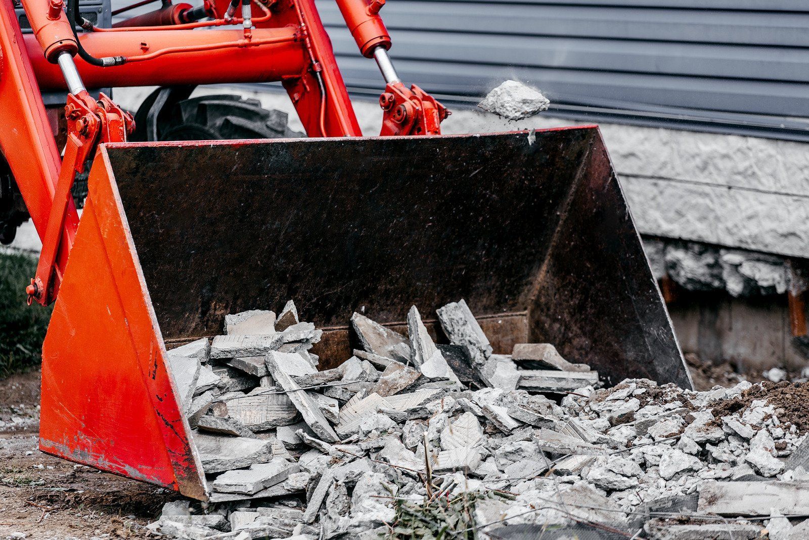 Red excavator bucket filled with gray debris outdoors.